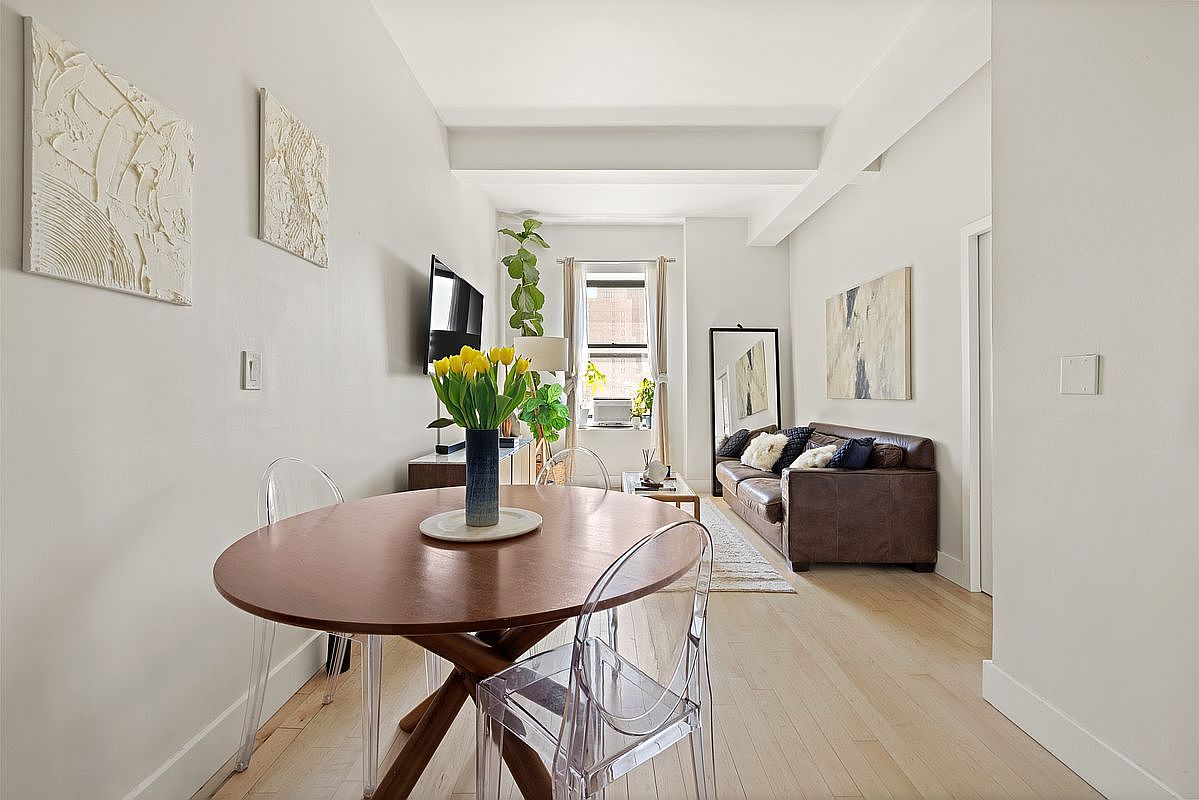 Open-concept dining area with round walnut table, ghost chairs, beamed ceiling overhead and brown leather sofa beyond near the east-facing window